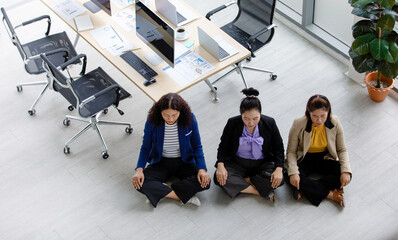 Top view shot of three Asian middle aged female businesswoman officer staffs in formal suit sitting squat quiet on floor close eyes meditating together at office working station table in company