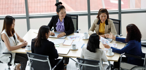 Asian middle aged female handicapped disabled businesswoman officer staff in formal business clothing sitting on wheelchair working with other workers with laptop computer in company office workspace
