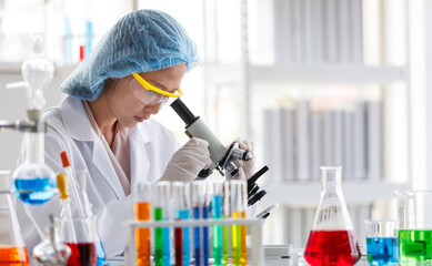 Fototapeta premium Close up shot of Asian female professional scientist in white lab coat safety glasses hygiene cap and rubber gloves using scientific microscope lens looking zooming sample on laboratory working desk