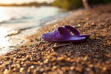 Close-up of rubber slippers on the shore of a lake or river made of small stones against a colorful sunset background. A beautiful place in nature for family holidays and swimming.