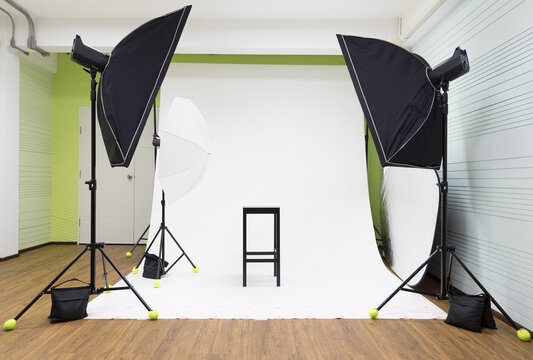 Empty nobody indoor room studio white photoshoot scene with professional photographer equipment set softbox strobe flash umbrella reflector light bulb on tripod stand with black chair ready for model