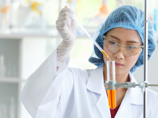Asian female professional scientist in white lab coat safety glasses hygiene cap and rubber gloves using dropper sucking orange liquid sample reagent from glass flask on laboratory analysis desk