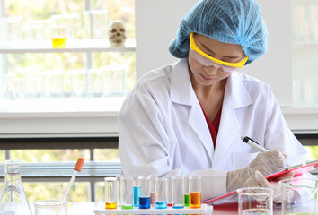 Asian female scientist in white lab coat safety goggles hygiene cap and rubber gloves writing result note on board when do science experiment on laboratory working desk with glass test tube beakers