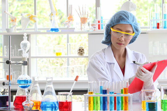 Asian Female Scientist In White Lab Coat Safety Goggles Hygiene Cap And Rubber Gloves Writing Result Note On Board When Do Science Experiment On Laboratory Working Desk With Glass Test Tube Beakers