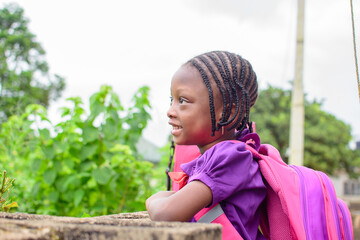 An African girl child in school uniform and with a backpack, resting her hands on a low fence