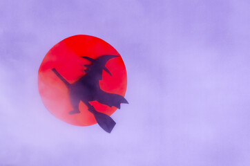 The shadow of a black witch flying on a broomstick in the clouds of fog against the background of the red moon. A mock-up of a colored invitation to a holiday.