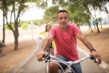 Fototapeta premium Portrait of active mature man riding bike in park with his wife in background. Positive senior man having weekend outdoors. Active seniors concept
