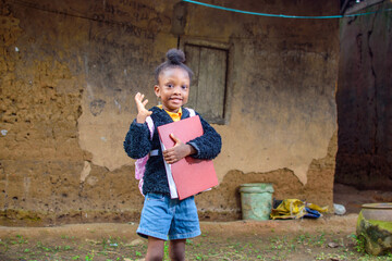 A happy little African girl child or student with pink school bag, holding and hugging her books outside a village mud house