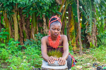 A pretty African lady or woman with beads on her head is grinding something with local grinding stone in a banana farm