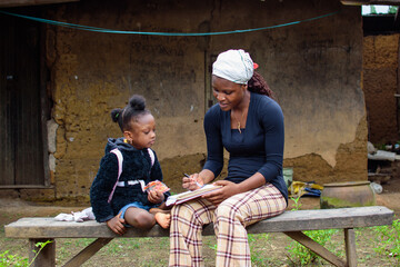 An African mother or teacher outside a village house, helping a girl child with her studies for...