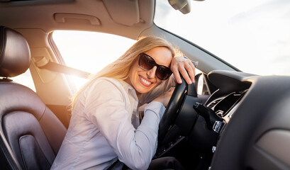 Smiling mature woman in sunglasses and blue shirt sitting on driver's seat inside modern car and leaning head on steering wheel. Business lady making stop during driving.