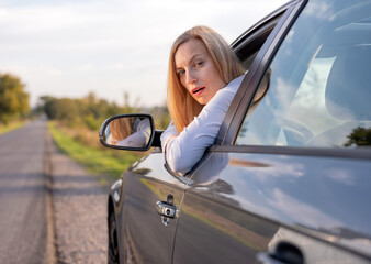 Mature woman with blond hair looking out of lowered glass of modern auto and feeling shocked. Problems with transport on road. Troubles during driving.