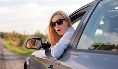 Fototapeta na wymiar Shocked mature woman in sunglasses sitting on driver's seat of modern car and looking out of lowered glass. aucasian blondie got in trouble while driving auto.