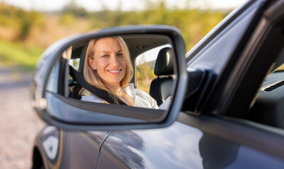 View in side mirror of beautiful caucasian woman with blonde hair sitting on drivers seat of modern car and fastening with safety belt. Concept of people and transportation.