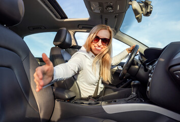 Fototapeta na wymiar Caucasian lady with blond hair giving lift to someone on road. Happy mature woman in sunglasses sitting on driver's seat and showing with hand on empty passenger seat.