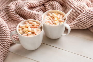 Cups of hot cocoa drink with marshmallows and plaid on light wooden background
