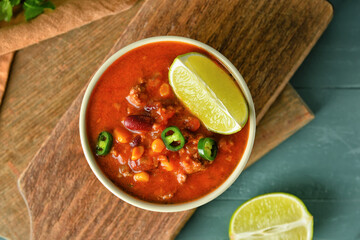 Bowl with tasty chili con carne and lime on color wooden background