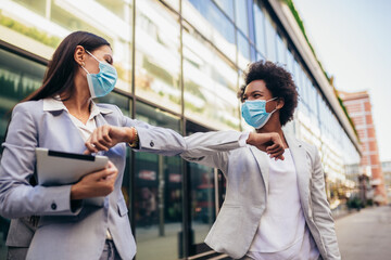 Business women with safety masks greeting with elbow bump in front of office building.