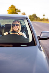 Happy mature woman in formal blue shirt and sunglasses, fastening with safety belt, driving modern...