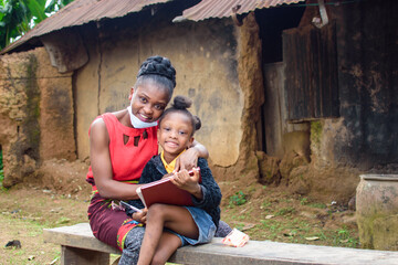 An African mother or teacher outside a village mud house with nose mask, helping a girl child with...