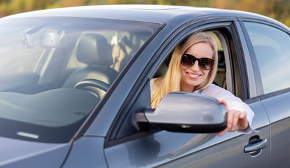 Fototapeta na wymiar Positive mature lady with blond hair in sunglasses and blue shirt adjusting side mirror before driving car. Concept of people and transport.
