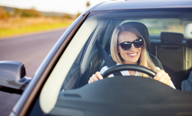 Happy mature woman in sunglasses and business wear enjoying driving her modern car. View of female...