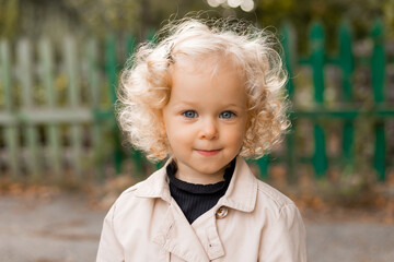 portrait of a cute little curly blonde girl with blue eyes in a white linen raincoat stands against...