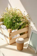 Box with chamomiles and cup of coffee on shelf near light wall