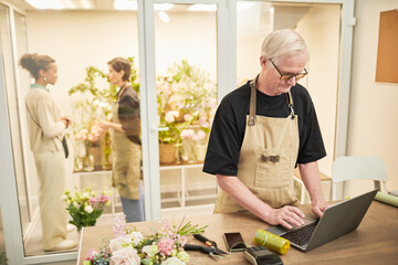 Portrait of male small business owner using laptop and wearing mask while managing flower shop,...
