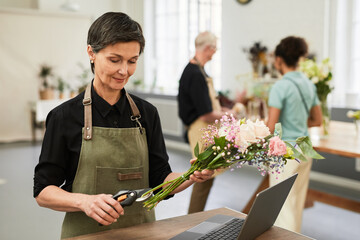 Portrait of elegant mature woman using arranging bouquets in flower shop while managing small...