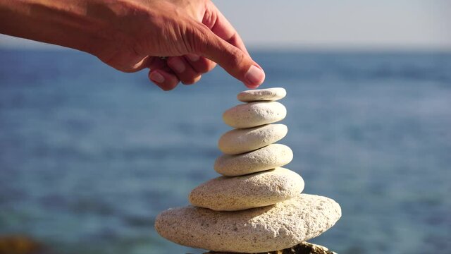 Man bilds stones pyramid on the seashore on a sunny day on the blue sea background. Happy holidays. Pebble beach, calm sea, travel destination. Concept of happy vacation on the sea, meditation, spa