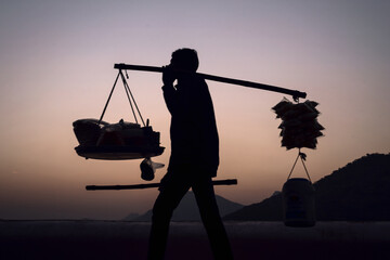 Silhouette of a street vendor carrying everything on his shoulder during sunset in Udaipur,...