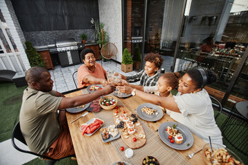 Fototapeta na wymiar Portrait of big African-American family clinking glasses while enjoying dinner together outdoors and celebrating