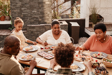 Warm toned portrait of happy African-American family sitting at table outdoors and enjoying dinner...