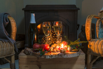 Christmas or winter holidays concept; home interior with two wicker chairs and rustic table on the wooden box; one bottle and two glasses of champagne, fruits and burning candles