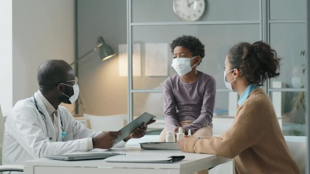 African American pediatrician in protective face mask giving health consultation to little boy and his mother while working in clinic during coronavirus pandemic