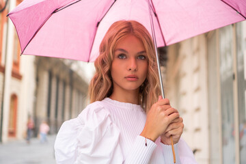 A portrait photo of a beautiful blond teenage girl with a pink umbrella on the street.
