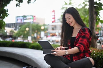 Young Asian woman sitting in a city park and using computer tablet.