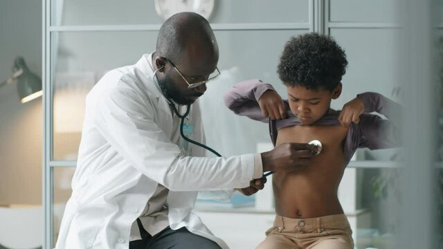 African American doctor in white coat using stethoscope while listening to heartbeat of little boy during medical checkup in clinic