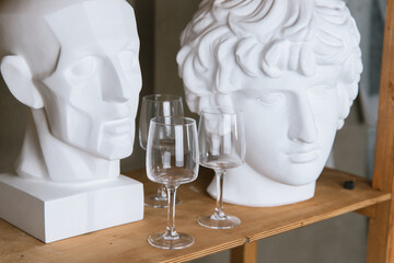 unusual empty wine glasses on a wooden shelf next to plaster heads, an art painting with wine glasses, bohemian wine glasses