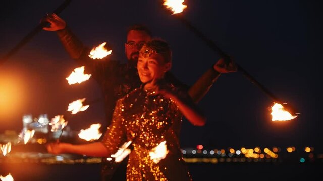 Fire show - smiling woman and man in a shiny clothes dancing with fire torches on the night beach