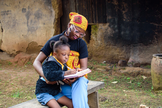 An African mother wearing glasses with a baby on her back, sits outside a village mud house, helping a girl wearing nose mask with studies for excellence in her school, career and education