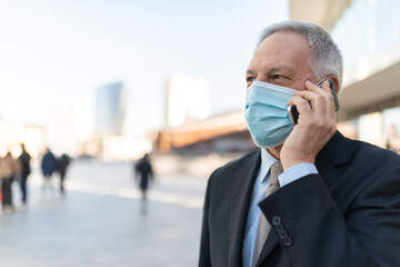 Covid coronavirus concept, masked elder man talking on his smartphone outdoor