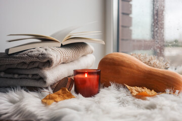 Burning candle, pumpkin, book, sweaters on the windowsill. Autumn mood.