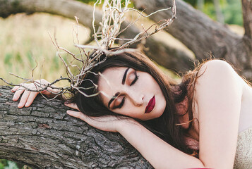 brunette girl Mavka Dryad Naiad forest nymph in a fairy tale in the forest among the trees in nature on her head a Hoop a wreath of branches dressed in a beige bra and silver fabric to the floor lying - obrazy, fototapety, plakaty