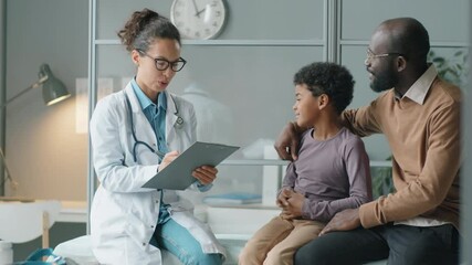 Beautiful female doctor taking notes and talking with little African American boy and his father while giving medical consultation in clinic - Powered by Adobe