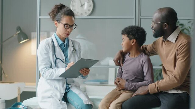 Beautiful female doctor taking notes and talking with little African American boy and his father while giving medical consultation in clinic