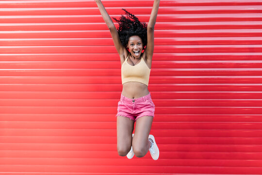 Carefree young woman with mouth open jumping in front of red shutter