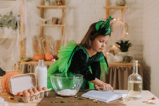 cute little girl in a green Halloween costume of a witch or fairy prepares a pumpkin pie, barm brack in a festive decorated kitchen. space for text. High quality photo