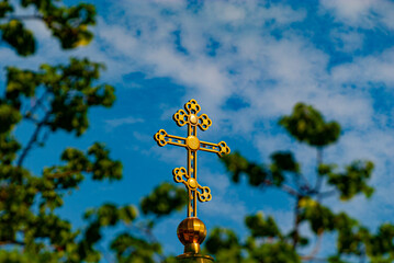 Fototapeta na wymiar a carved gilded shiny Orthodox cross is visible through the branches of trees against the background of a blue sky with light clouds
